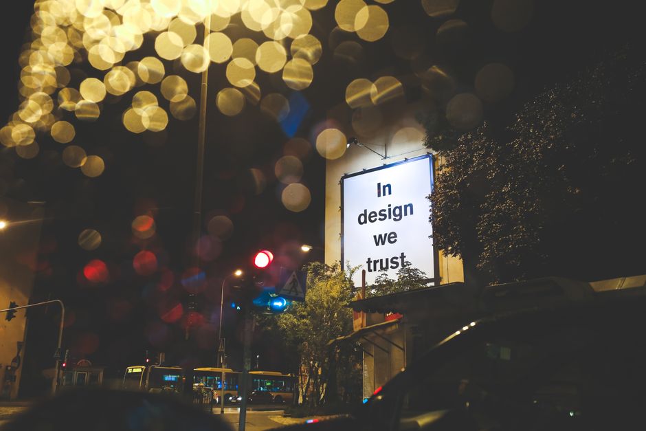 A white billboard reading “In design we trust” beside a street at night